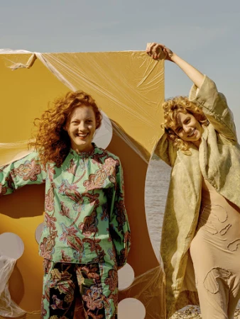 Two adult females pose outdoors next to a large yellow wrapped cube under a sunny sky. One wears patterned separates and the other, neutral layers.