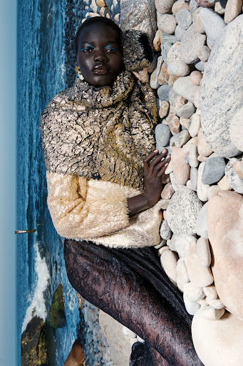 A woman with dark skin and blue eye makeup poses in a gold and black patterned jacket with water and rocks in the background.