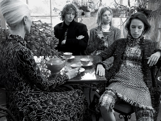 A group of four people sitting at a table with teacups and a teapot in a bright indoor setting, all dressed in patterned clothing.