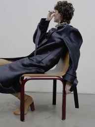 A person with short curly hair wearing a dark coat and tan boots sits backward on a red-framed wooden chair against a plain background.
