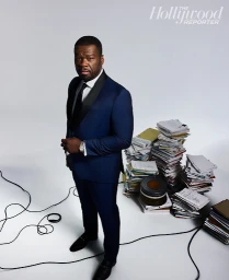 50 Cent wearing a blue tuxedo suit standing beside stacked piles of paper documents against a white studio background.