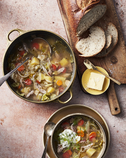 Overhead shot of chicken soup in a pot and a bowl, served with slices of bread and butter on a wooden board.