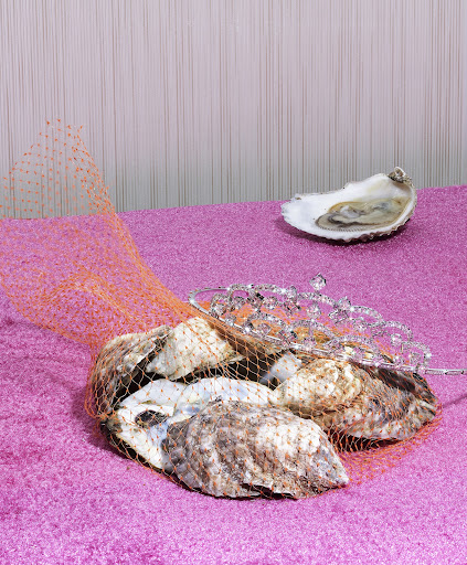 Still life of oysters and a tiara on a purple surface, draped with orange netting against a striped beige background.