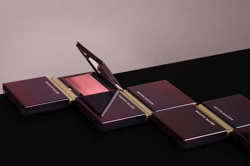 Compact makeup palette by Kevyn Aucoin with a mirror and visible blush in a beauty editorial. Products lined up on a black surface.