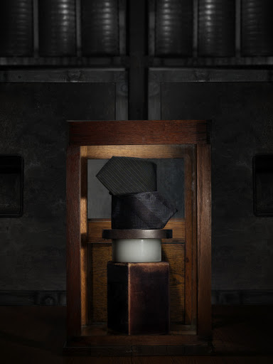 Still life shot of stacked ties in a wooden frame atop white and dark brown pedestals, set against a dark, industrial background.