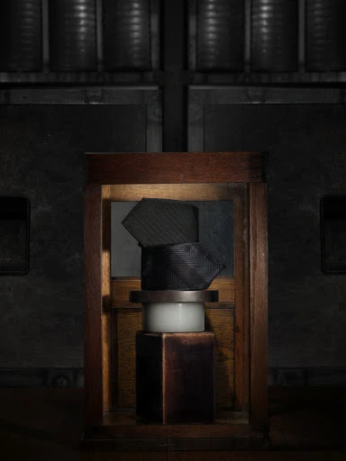Still life shot of stacked ties in a wooden frame atop white and dark brown pedestals, set against a dark, industrial background.