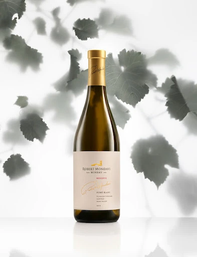 Elegant bottle of Robert Mondavi Reserve white wine on a white surface with soft shadows and leaf pattern in the background.
