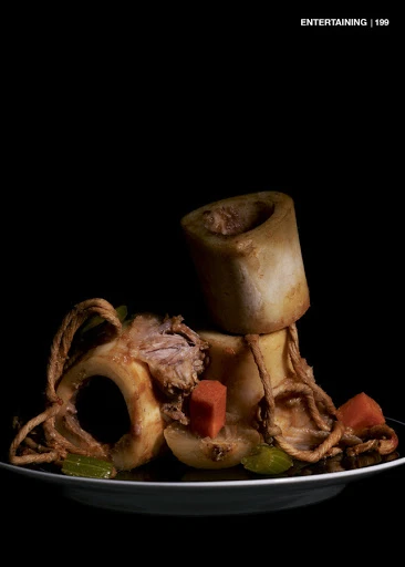 Studio shot of beef shank dish featuring cooked beef shanks, broth, carrots, and celery on a dark plate against a black background.