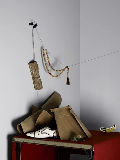 A still life featuring a white shoe, brown paper, beaded necklace, and other props against a gray wall, creating an artful composition.