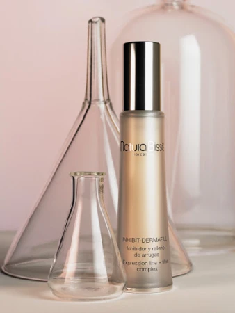 Natura Bisse Inhibit-Dermafill skincare bottle surrounded by glass laboratory glassware on a soft pink surface