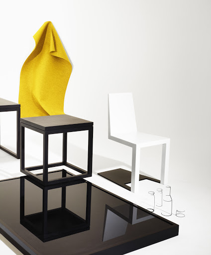 Minimalist studio shot featuring geometric furniture: black cube tables, a white chair, a glossy black table, and a mustard-yellow cloth.