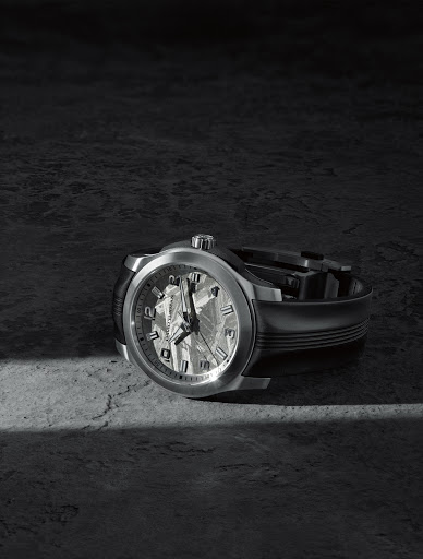 Elevated view of a luxury wristwatch with a silver band and intricate face, resting on a textured gray surface in a studio setting.