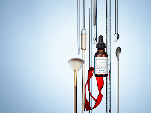 Beauty serum bottle with dropper, makeup brush, lab glassware, and red glasses on a light blue background in a studio shot.