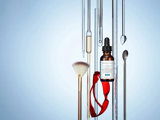 Beauty serum bottle with dropper, makeup brush, lab glassware, and red glasses on a light blue background in a studio shot.