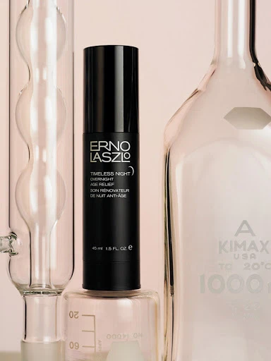 Erno Laszlo skincare product, Timeless Night Age Relief, in black pump bottle on lab glassware, soft pink backdrop, beauty product.