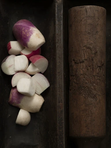 Overhead close-up of chopped turnips and radishes in a dark tray next to a wooden rolling pin.