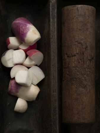 Overhead close-up of chopped turnips and radishes in a dark tray next to a wooden rolling pin.