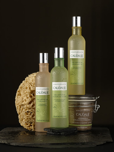 Caudalie Vinothérapie products featuring shower gel, scrub, and sponge against a dark background and wet slate surface.
