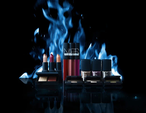 Product shot of NARS brand cosmetics, including lipsticks, glosses, and eyeshadow compacts, against a dramatic blue flame background.