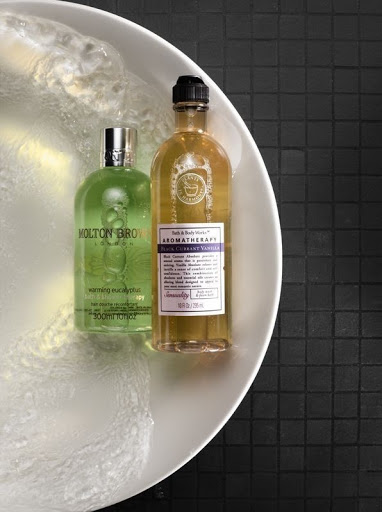 Still life shot of Molton Brown and Bath & Body Works Aromatherapy bottles in a white bowl with water against a black tiled background.