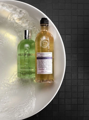 Still life shot of Molton Brown and Bath & Body Works Aromatherapy bottles in a white bowl with water against a black tiled background.