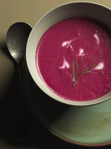 Overhead shot of vibrant pink beet soup garnished with cream and dill in a white bowl, placed on a green plate with a silver spoon.
