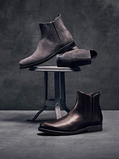 Studio shot of men's ankle boots in brown leather and grey suede displayed on a metal stool against a dark grey textured background.