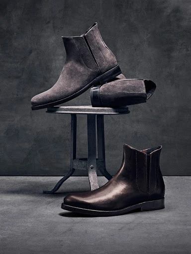 Studio shot of men's ankle boots in brown leather and grey suede displayed on a metal stool against a dark grey textured background.