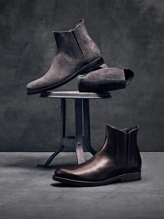 Studio shot of men's ankle boots in brown leather and grey suede displayed on a metal stool against a dark grey textured background.