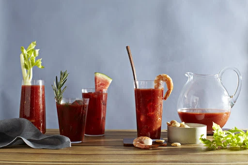 Bloody Mary cocktails arranged with garnishes in glasses and a clear pitcher on a wooden table against a light blue backdrop.
