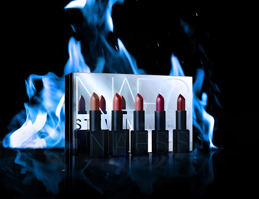 Cosmetic product shot: NARS lipstick collection displayed against blue flame effect on a reflective black surface with logo text visible.