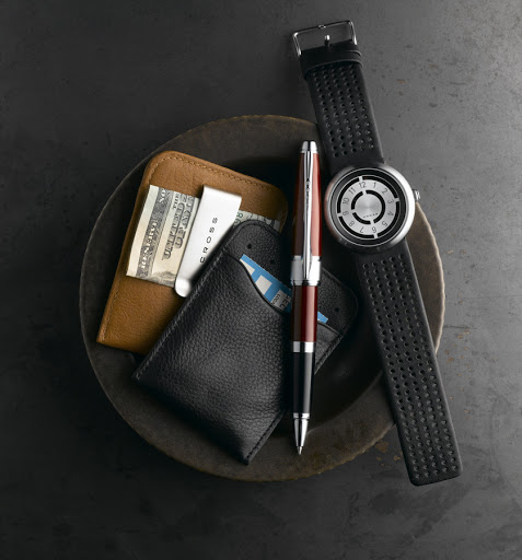 Overhead still life of leather wallets, a fountain pen, and a black watch in a dark bowl on a textured grey surface