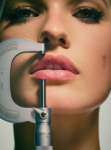 Close-up portrait of a fair-skinned adult female's face with blue eyes and pink lips, a metal micrometer calipers measuring her lips.