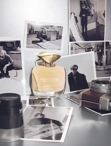 Still life featuring perfume bottle with gold cap surrounded by black and white portrait photographs on shiny metallic surface.