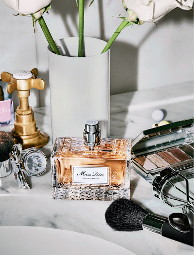 Crystal bottle of Miss Dior perfume surrounded by makeup brushes, eye shadow palette, and white roses against a marble backdrop.