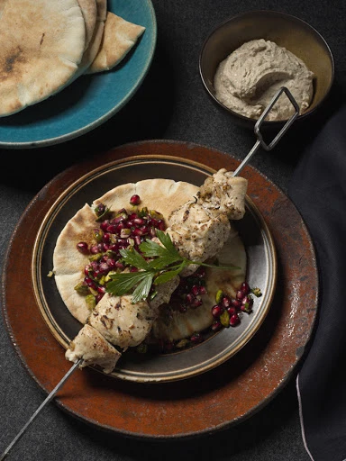 Overhead shot of chicken kebab skewer on pita bread with pomegranate seeds and parsley, served with flatbread and dip.