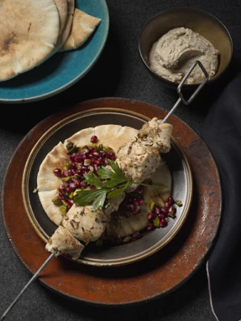 Overhead shot of chicken kebab skewer on pita bread with pomegranate seeds and parsley, served with flatbread and dip.