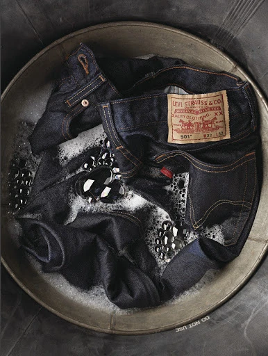 Close-up of a metal wash tub with Levi Strauss & Co. denim jeans soaking in soapy water with bubbles. Brand label is visible.