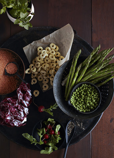 Overhead shot of uncooked pasta rings, asparagus spears, peas, radicchio, and radishes arranged on a dark wooden table.
