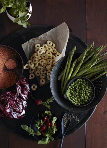 Overhead shot of uncooked pasta rings, asparagus spears, peas, radicchio, and radishes arranged on a dark wooden table.