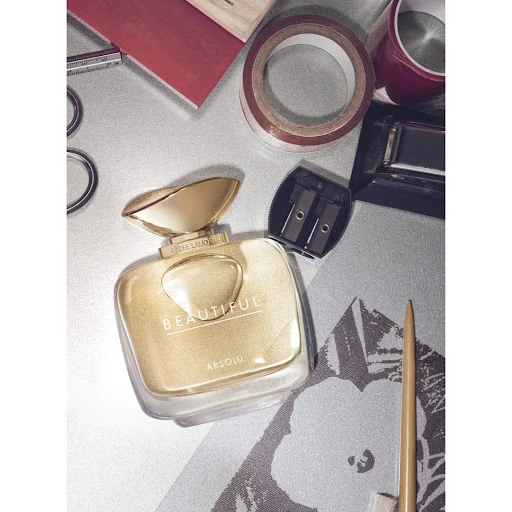 Elegant square perfume bottle of Estee Lauder's Beautiful Absolu with golden lid surrounded by makeup tools on a silver surface.