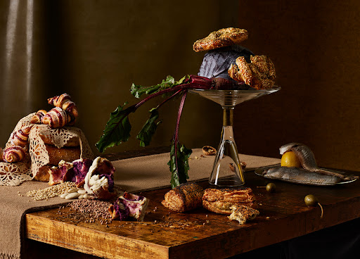 Artistic food still life on a rustic wooden table features pastries, grains, beetroot, and fish, creating a visually rich composition.