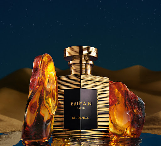 Balmain Paris Sel D'Ambre perfume bottle with gold accents, amber stones, set against a blue and tan background evoking a desert landscape.