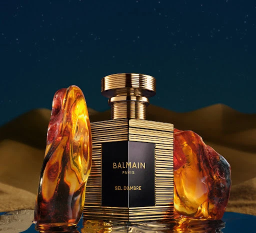 Balmain Paris Sel D'Ambre perfume bottle with gold accents, amber stones, set against a blue and tan background evoking a desert landscape.
