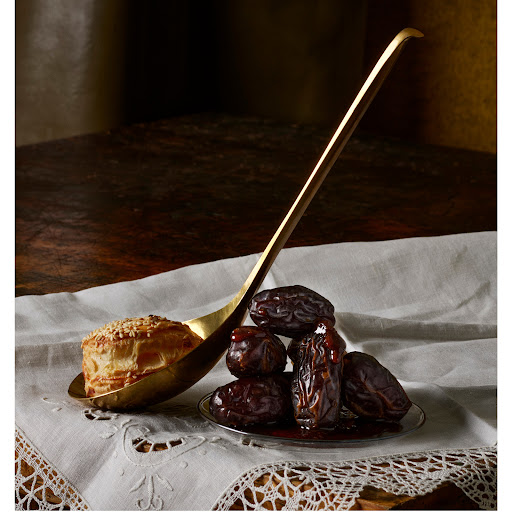 Still life shot of a pastry on a gold spoon, next to a pile of dates on a silver plate, set on a white, patterned tablecloth.