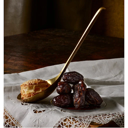 Still life shot of a pastry on a gold spoon, next to a pile of dates on a silver plate, set on a white, patterned tablecloth.
