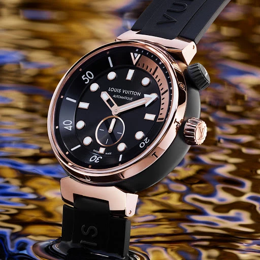 Luxury black and rose gold Louis Vuitton Tambour watch with black rubber strap on a reflective gold and blue surface, close-up.