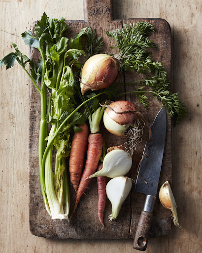 Overhead shot of fresh celery, carrots, and onions arranged on a wooden cutting board with a knife, creating a rustic food still life.