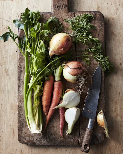 Overhead shot of fresh celery, carrots, and onions arranged on a wooden cutting board with a knife, creating a rustic food still life.