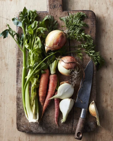 Overhead shot of fresh celery, carrots, and onions arranged on a wooden cutting board with a knife, creating a rustic food still life.